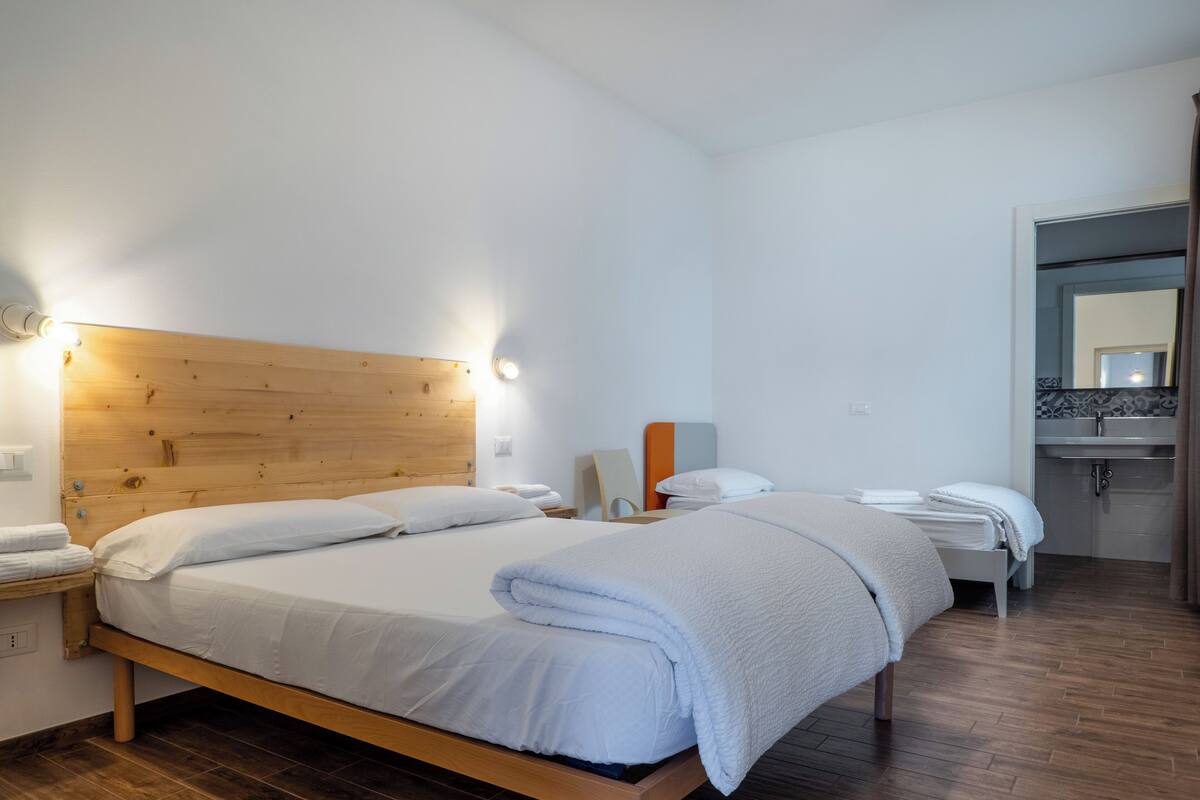 A hotel room with a wooden headboard, two beds, a nightstand, a bedside table, a mirror, and a bathroom with a sink and mirror.