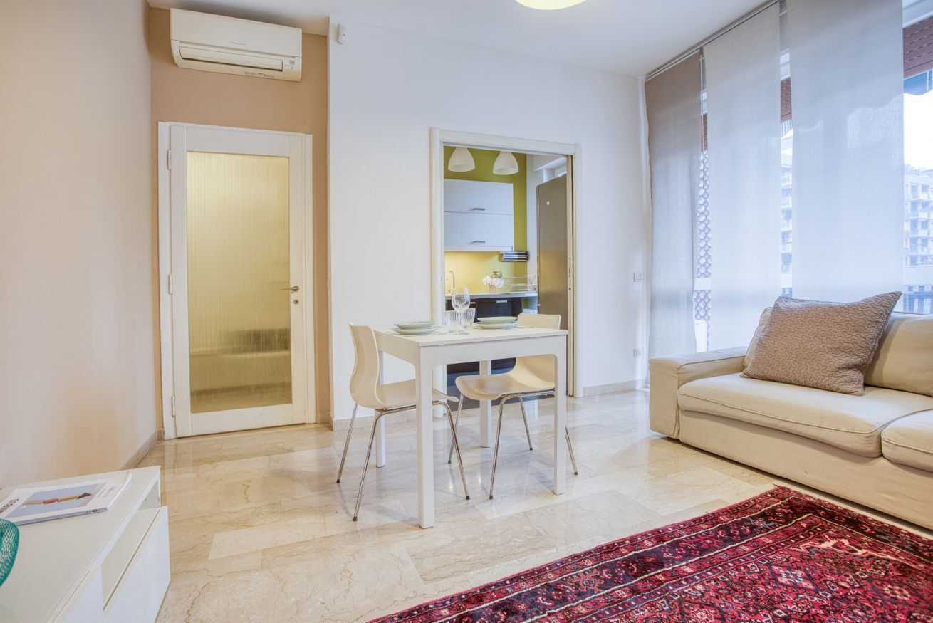 Ein modernes Wohnzimmer mit einem beigen Sofa, einem Esstisch mit Stühlen, einem roten Teppich mit Muster und einer durch eine offene Tür einsehbaren Küche.
