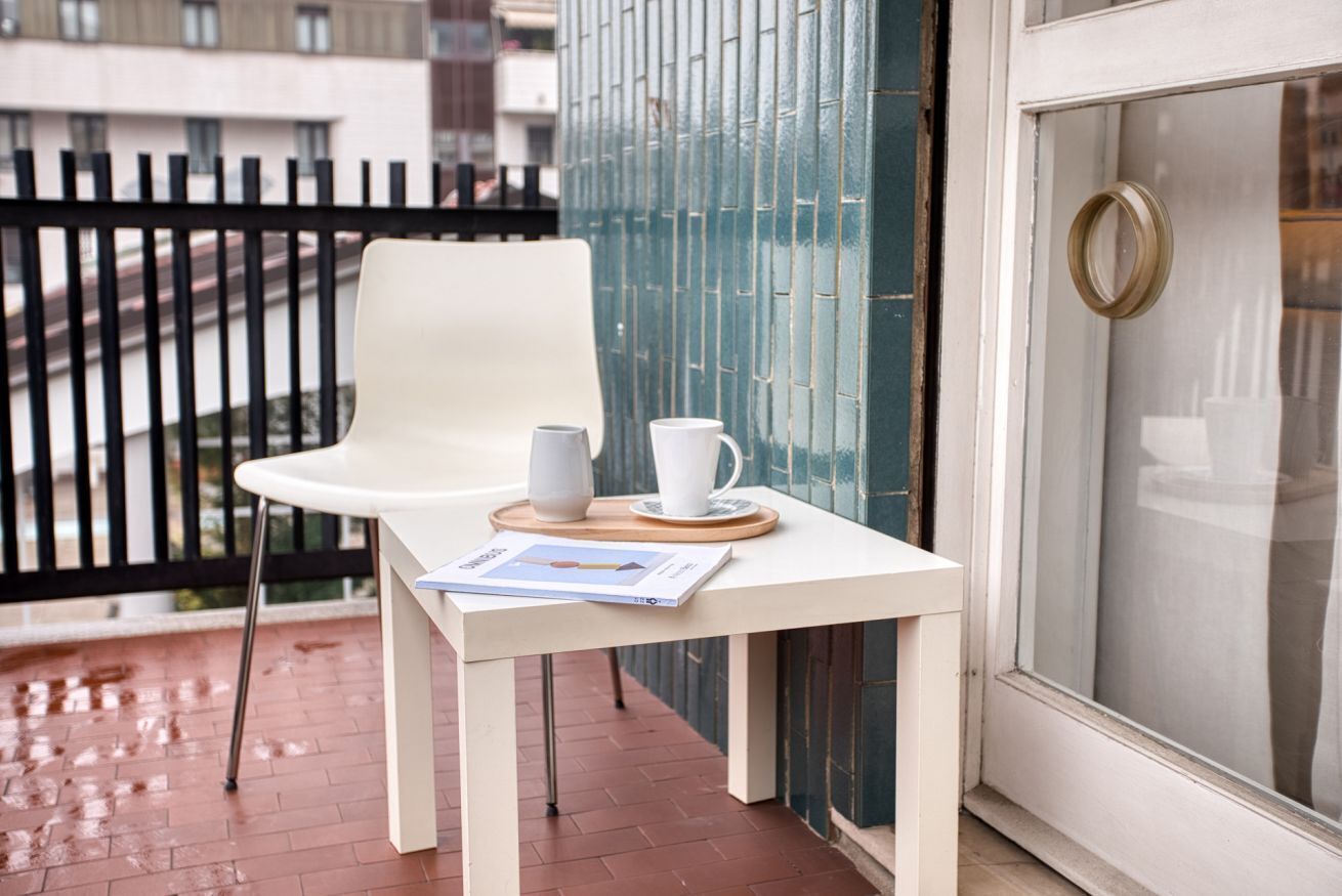 Ein weißer Tisch mit einem Stuhl, einer weißen Tasse und einer Zeitschrift auf einem Balkon mit Glastür und Metallgeländer.