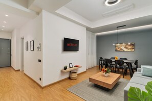 A modern living room with a wooden coffee table, a TV mounted on the wall, a dining area with a table and chairs, and a grey sofa.