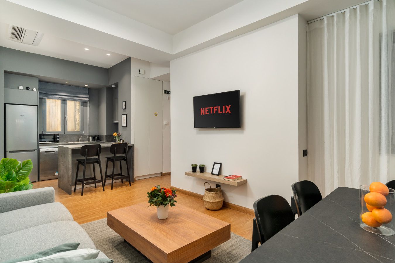 Un salon moderne comprenant une cuisine, un coin repas et un téléviseur fixé au mur, sur lequel on peut lire l’inscription « NETFLIX ».