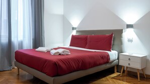 A neatly made bed with red bedding and white pillows, a bedside table with a book, and a modern wall-mounted light fixture.