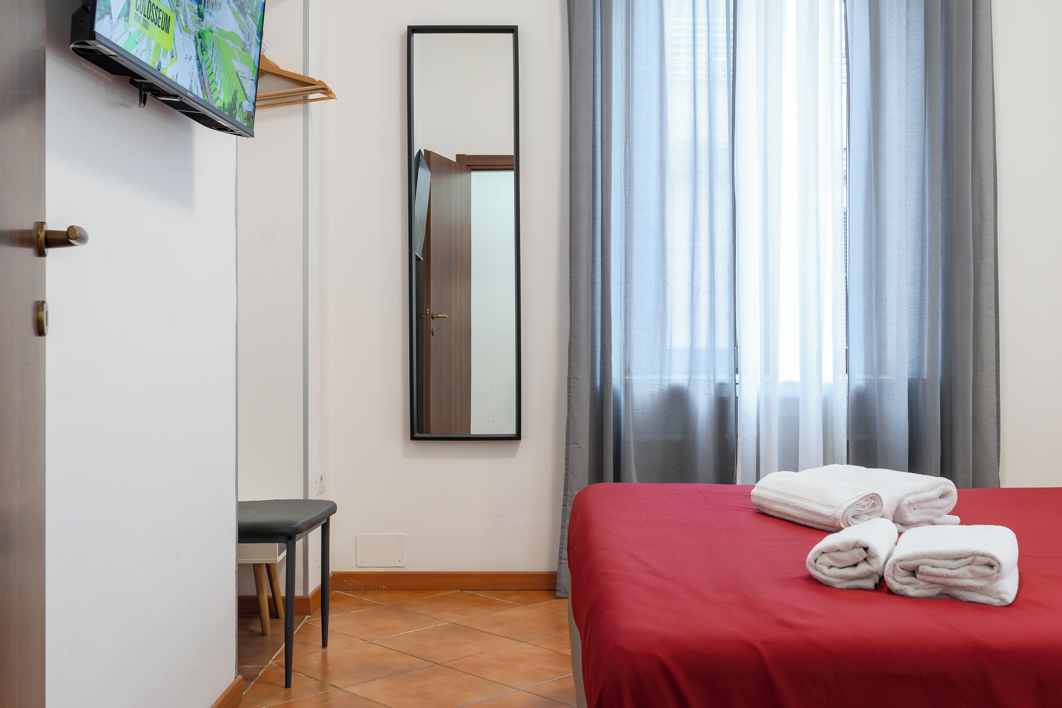 A hotel room with a bed, a TV mounted on the wall, a mirror, and a window with curtains.
