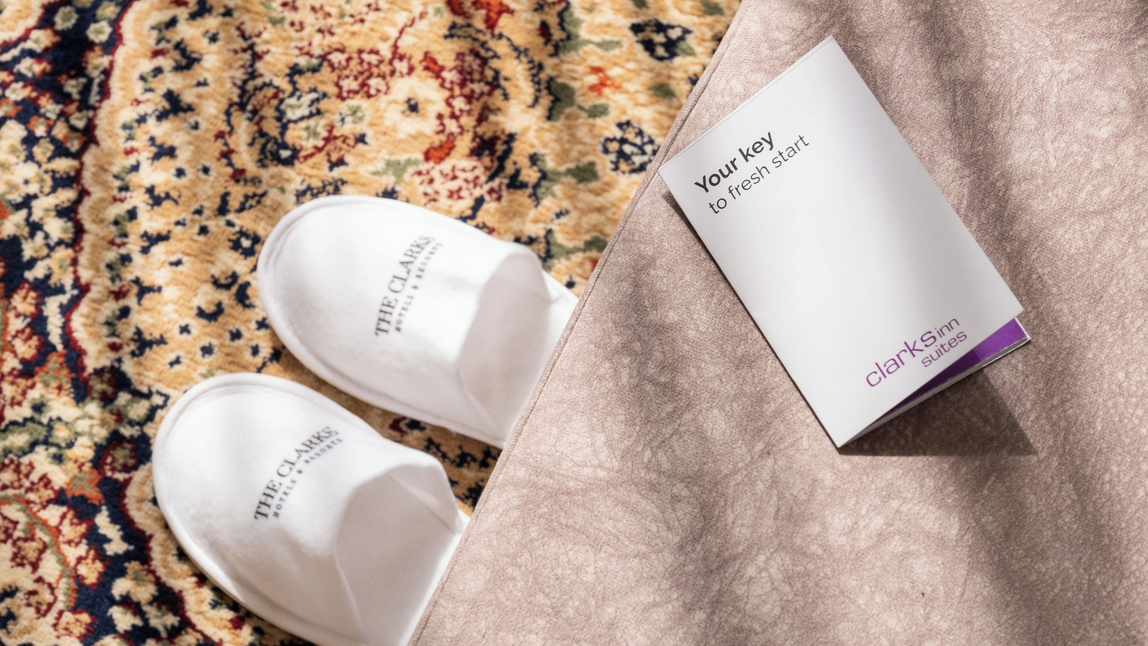 A pair of white slippers with "THE CLARKS" branding, a white card with "Your key to fresh start" text, and a "CLARKS inn suites" label on a textured fabric surface.