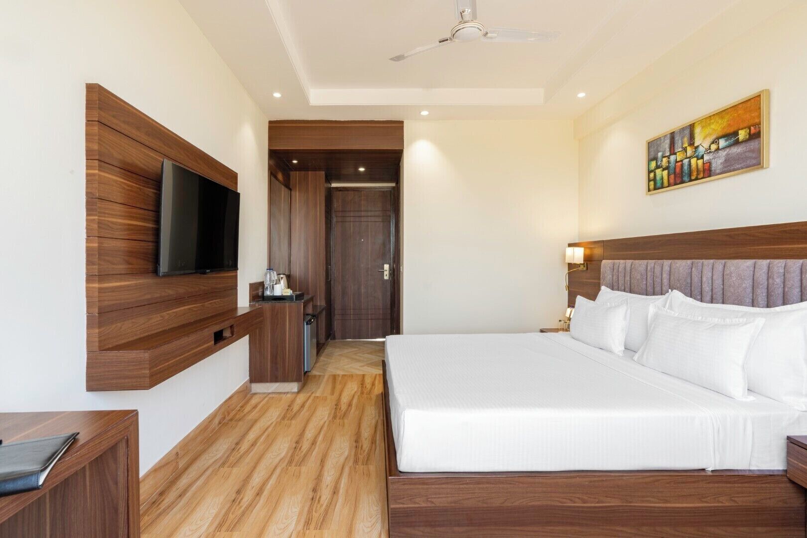 A hotel room with a large bed, a TV mounted on the wall, a desk with a chair, and a painting on the wall.