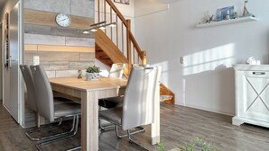 Ein Essbereich mit einem Holztisch, grau gepolsterten Stühlen, einer Wanduhr und einer Treppe mit Holzbalken.