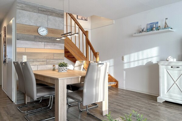 A dining area with a wooden table, grey upholstered chairs, a clock on the wall, and a staircase with wooden railings.