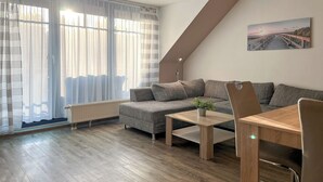 Ein modernes Wohnzimmer mit einem grauen Sofa, einem Holztisch und einem Esstisch mit Stühlen. An der Wand hängt ein Gemälde, das einen Sonnenuntergang über einer Brücke zeigt.