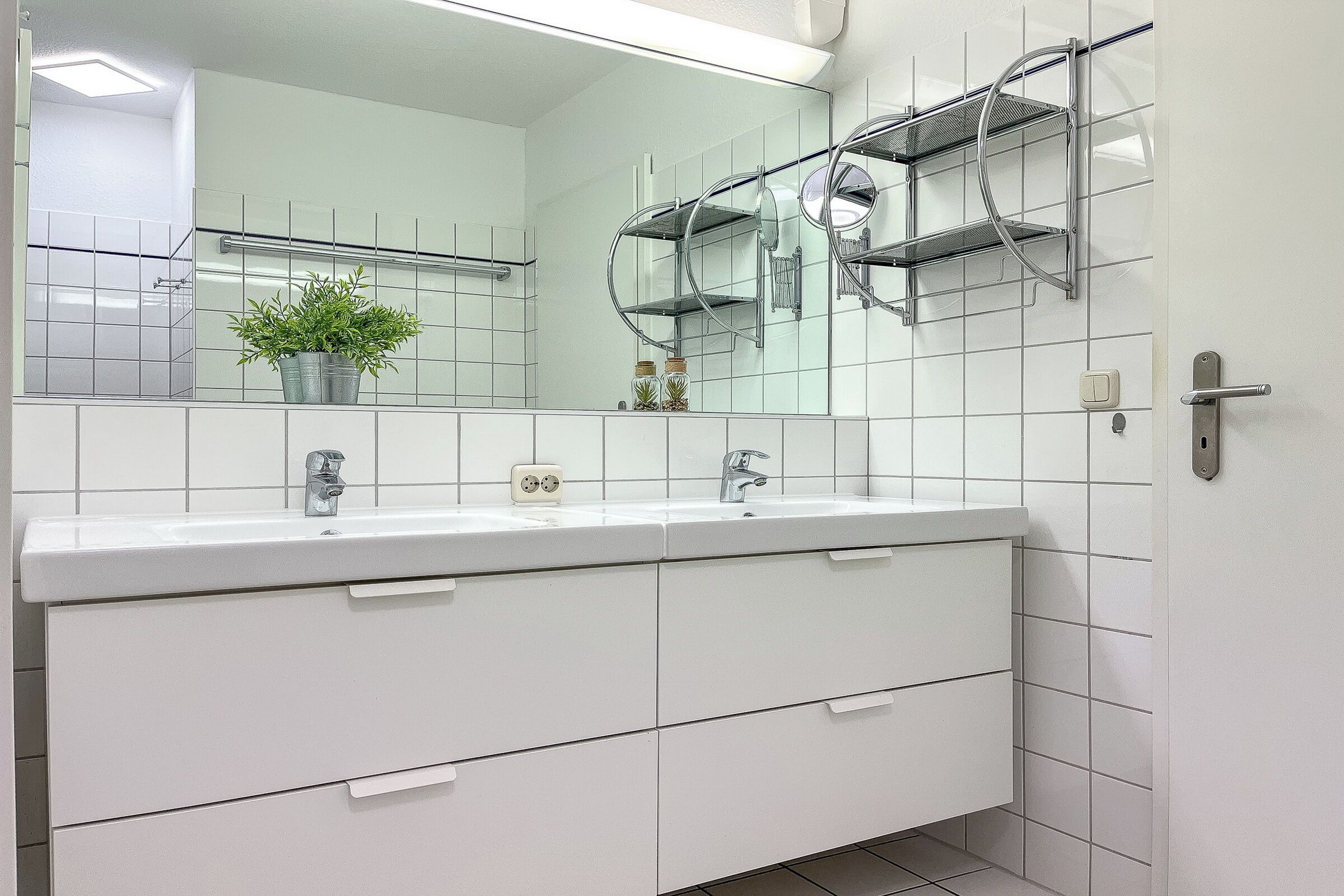 Een moderne badkamer met een witte wastafel, twee wasbakken, een grote spiegel en een kamerplant.
