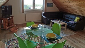 Ein gemütliches Loft-Zimmer mit einem Glastisch, grünen Stühlen, einer schwarzen Ledercouch und einem Fernseher auf einem Holzbodenregal.