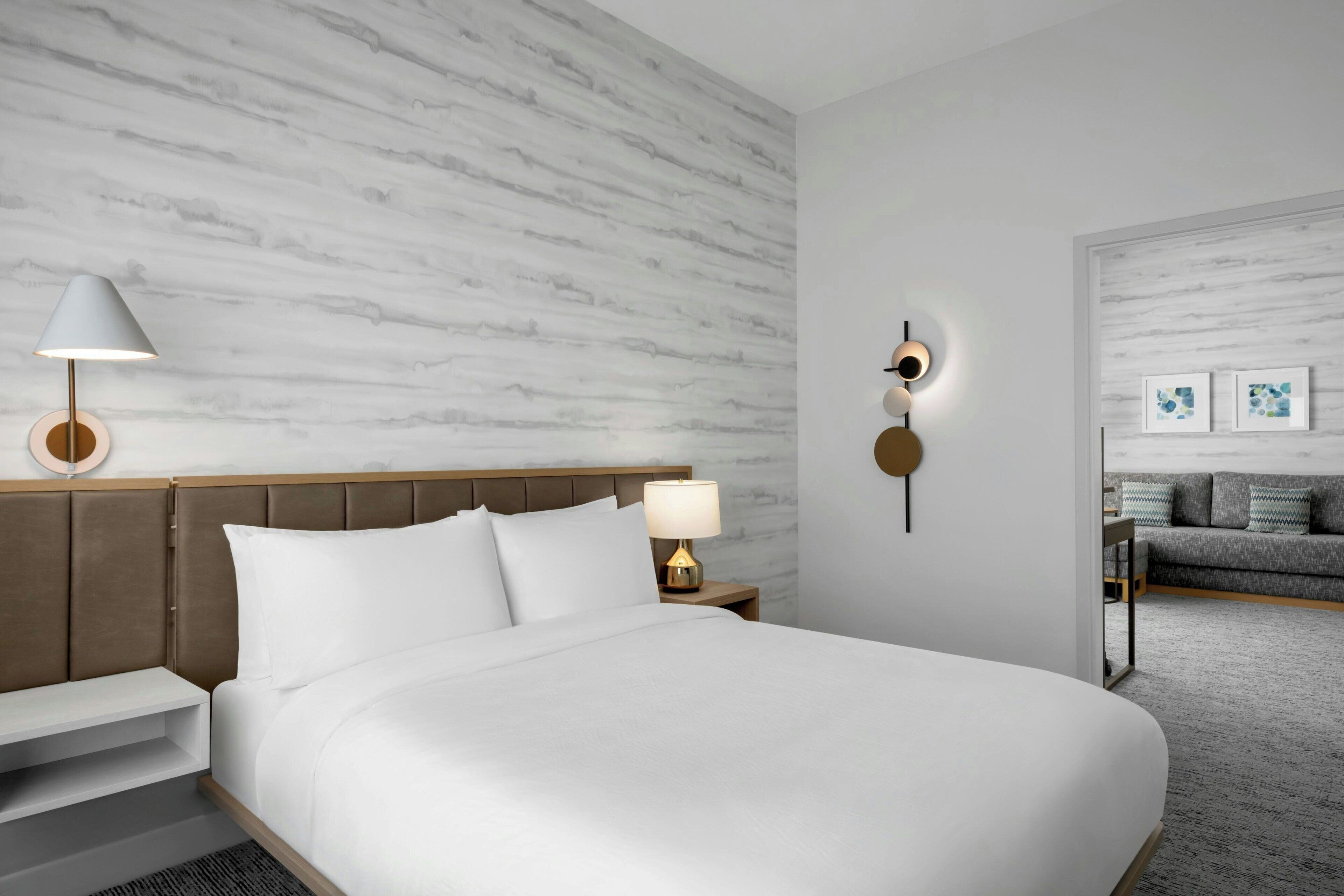 Habitación de hotel con una cama grande, una mesita de noche con lámpara, una lámpara de pared y un sofá en la habitación contigua.