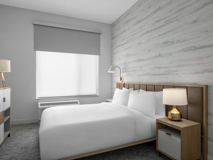 A modern hotel room with a large bed, a nightstand with a lamp, a dresser, and a television on the left.