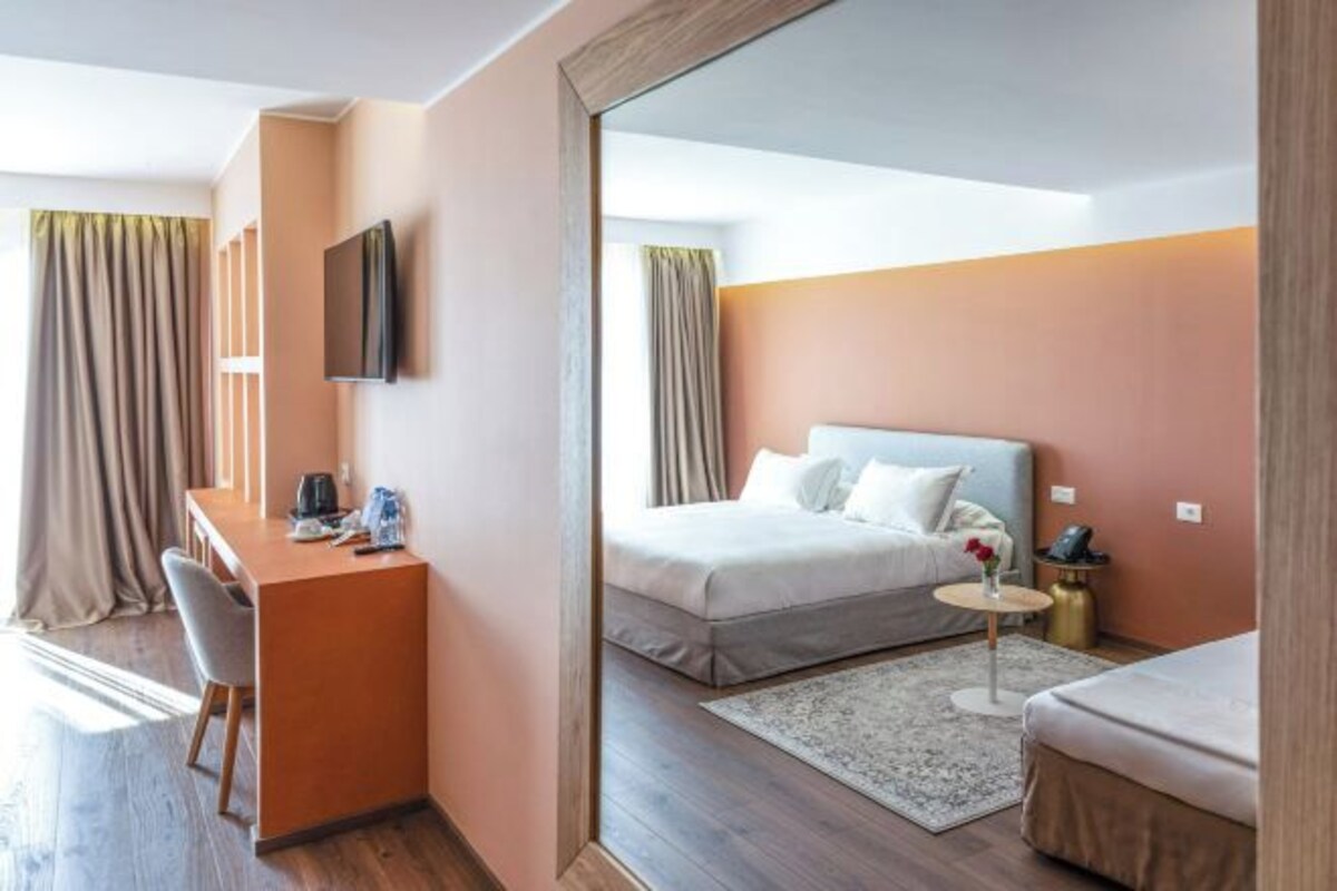 A hotel room with a bed, a desk, a chair, a small table with a vase, and a mirror.