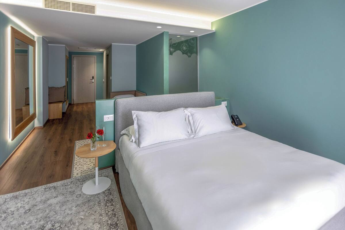 Quarto de hotel moderno com uma cama grande, uma mesinha de cabeceira com luminária, uma pequena mesa redonda com um vaso de flores e um espelho na parede.