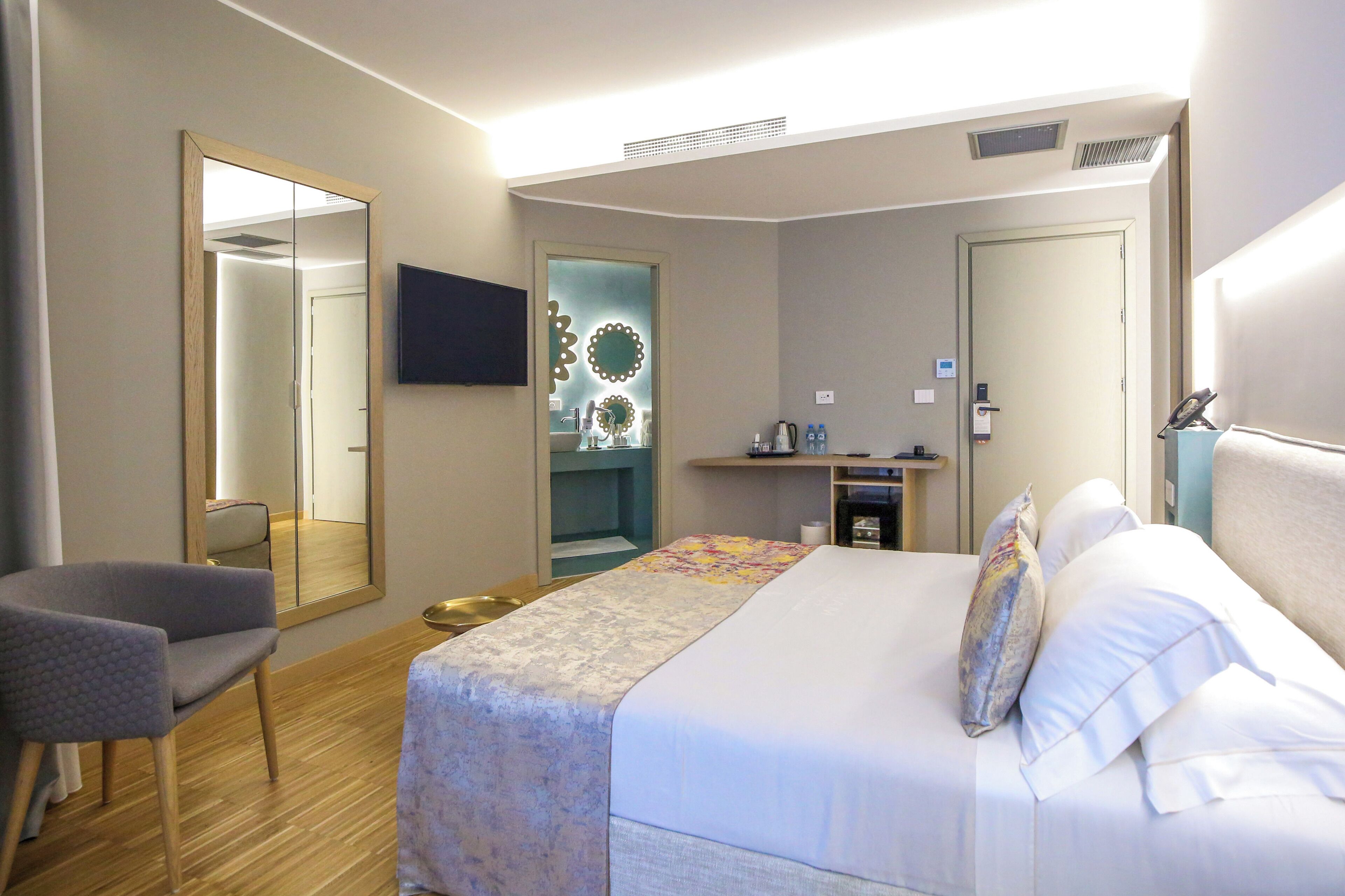 Habitación de hotel con cama, una silla, televisor, espejo, baño y una mesita de noche con artículos de aseo.