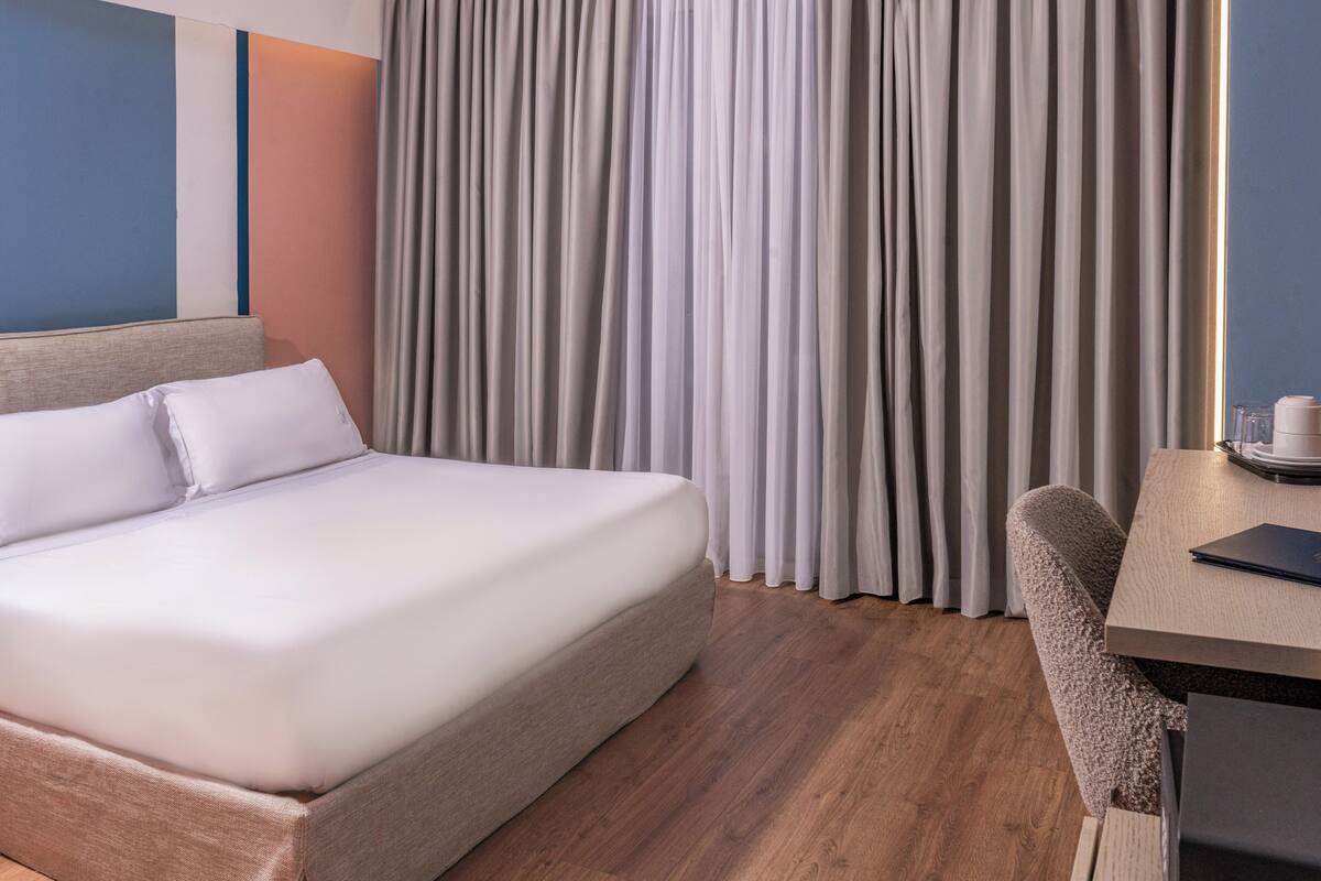 Quarto de hotel com cama, escrivaninha e janela com cortinas.
