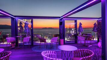 An outdoor terrace with tables and chairs, large windows, and a view of the sunset over the water.