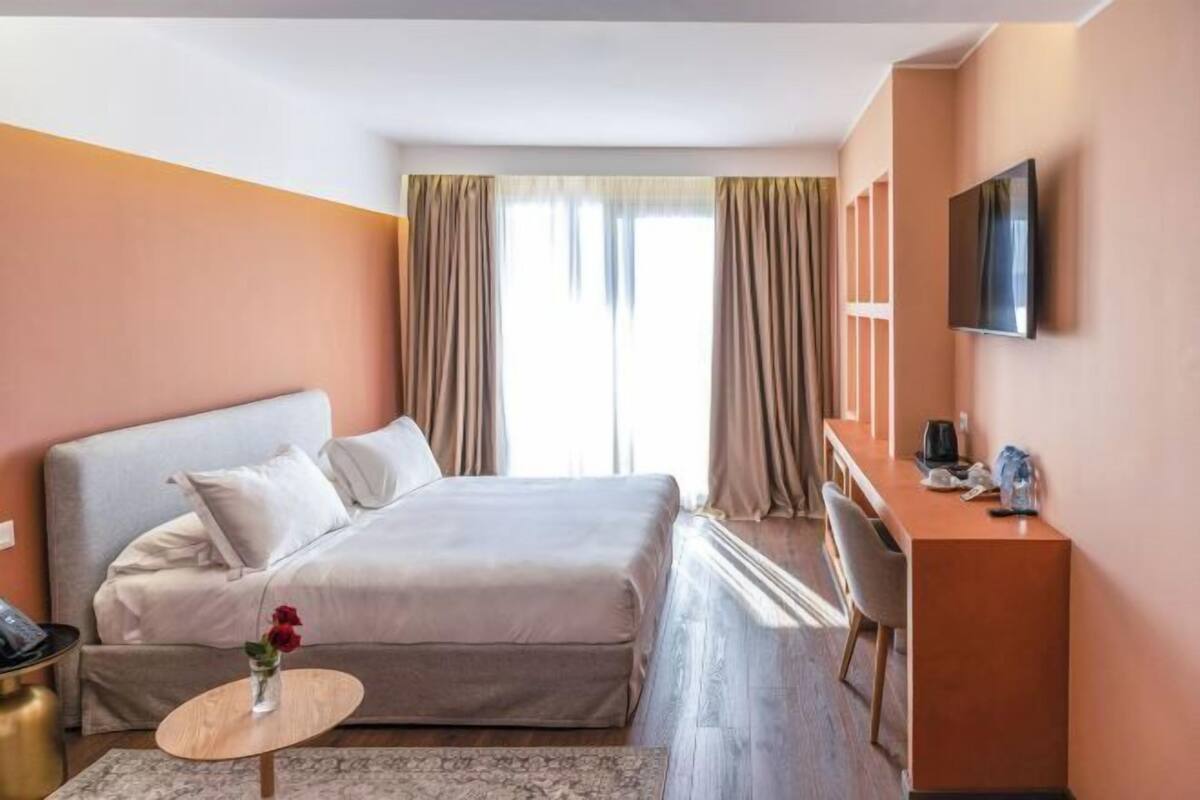 Quarto de hotel com cama, secretária, televisão e janela com cortinas.