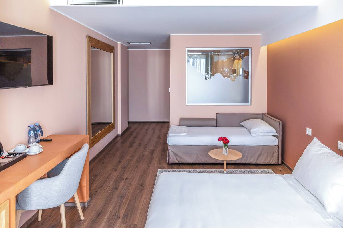 Quarto de hotel com escrivaninha de madeira, uma cama, mesa redonda pequena com um vaso de flores e um espelho grande na parede.