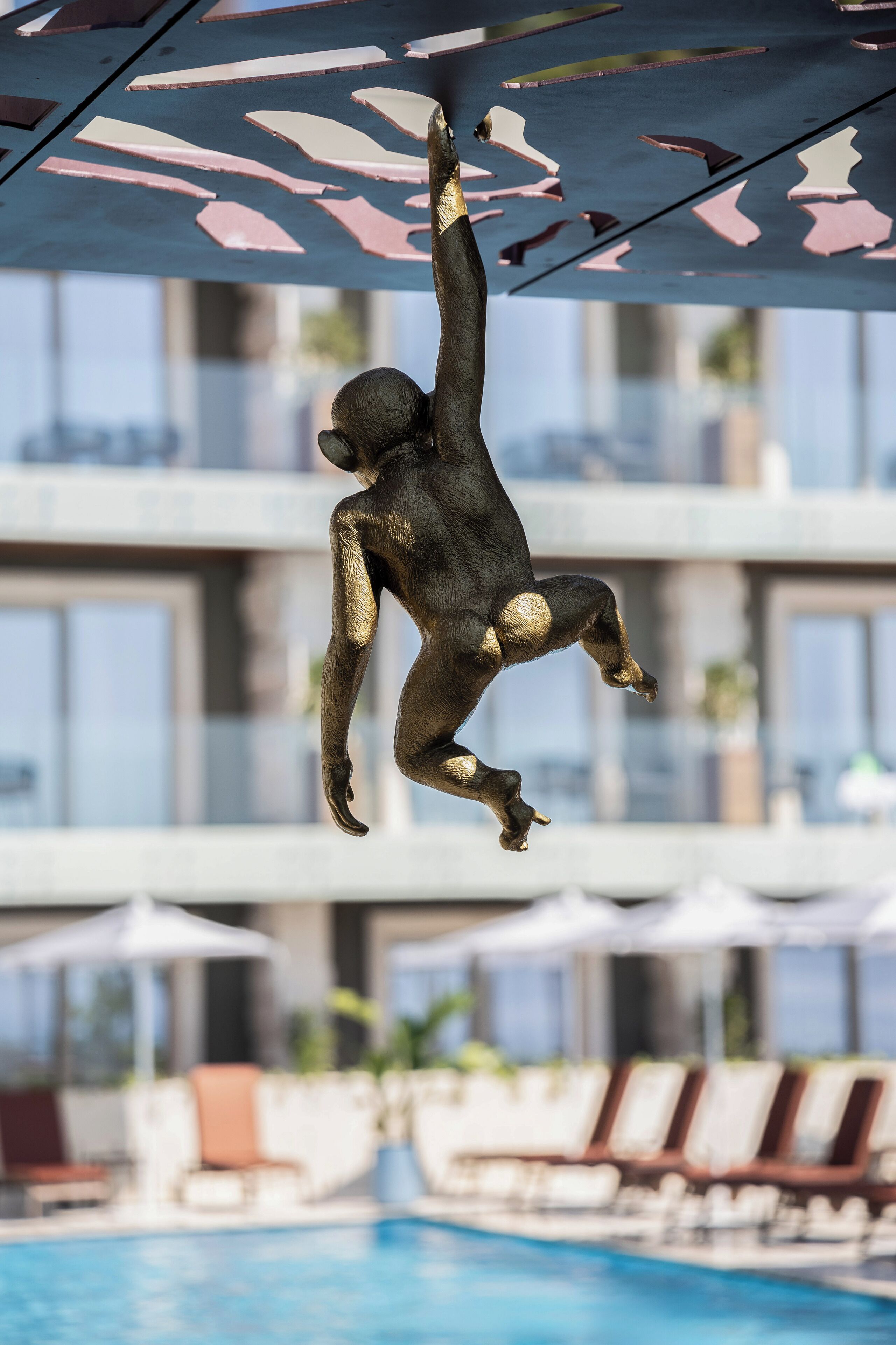 A monkey sculpture hanging from a ceiling with a pool and lounge chairs in the background.