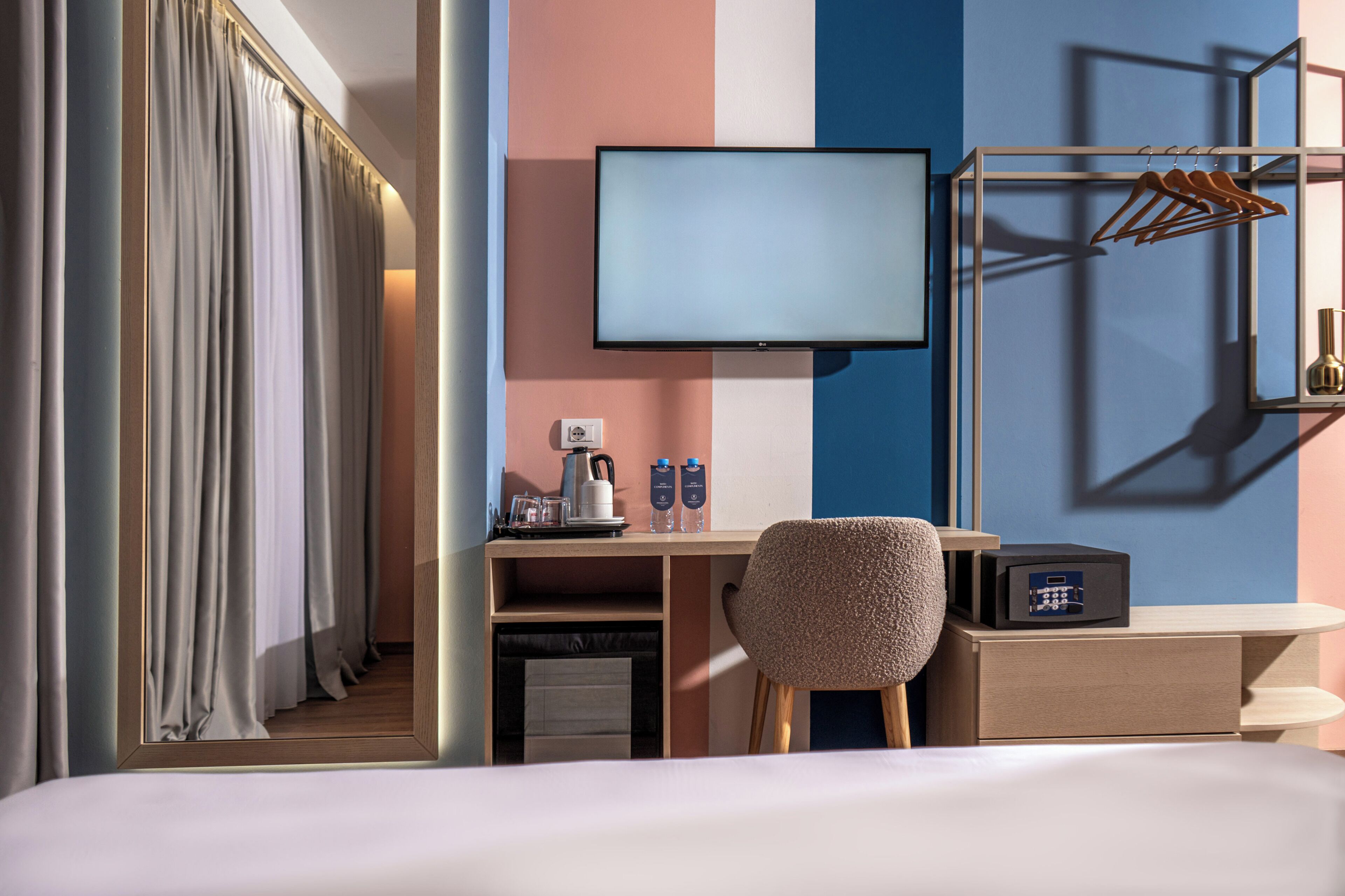 Une chambre d’hôtel comprenant un lit, un bureau avec un ordinateur, une chaise, un téléviseur, un porte-manteau et un mini-bar.