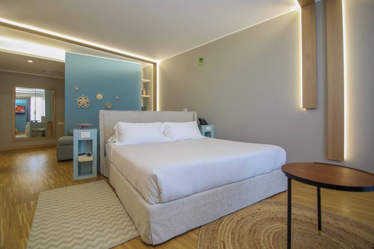 Quarto de hotel moderno com uma cama grande, mesas de cabeceira, uma pequena mesa redonda e um espelho.