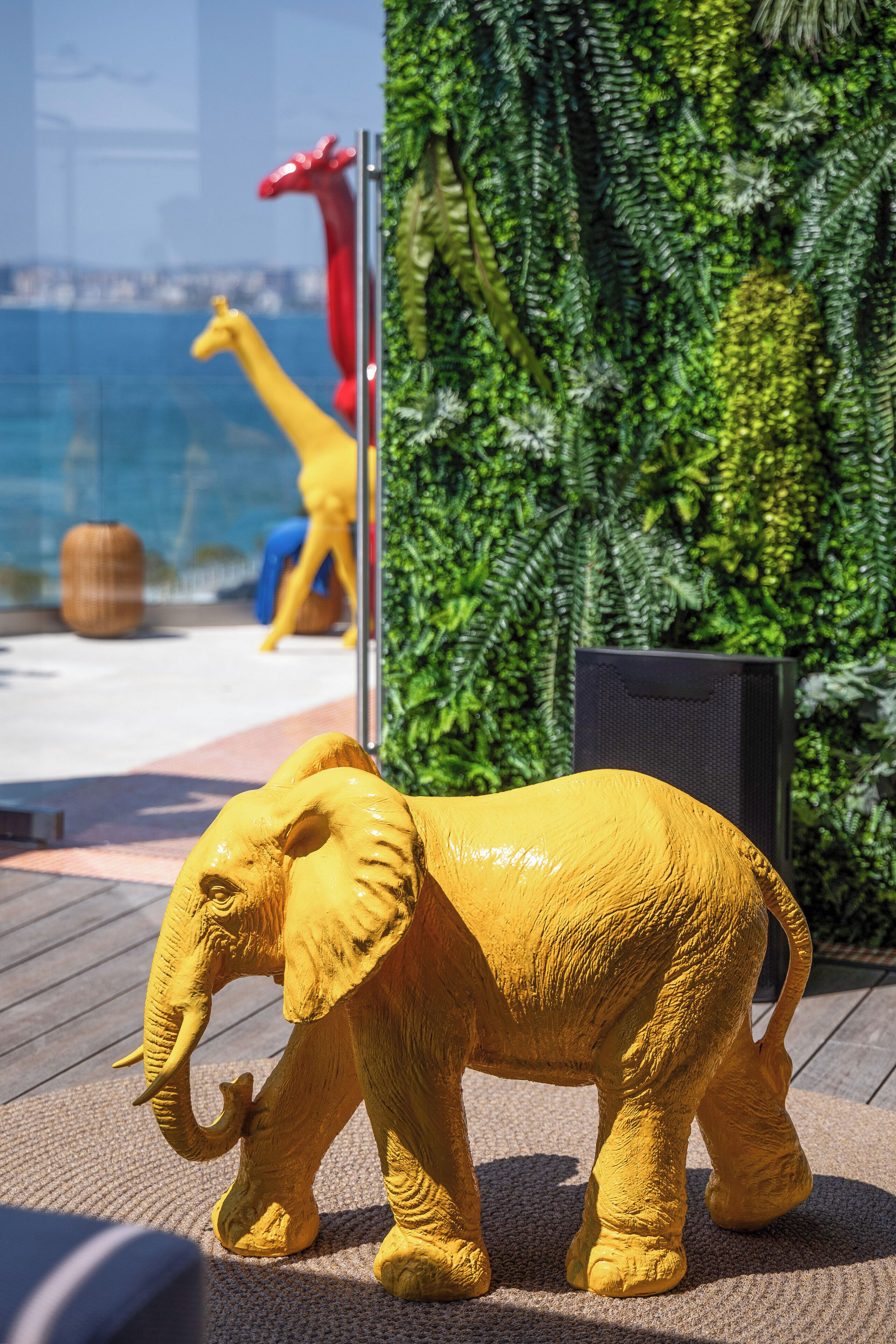 A golden elephant sculpture on a wooden deck with a backdrop of green foliage and a glass wall reflecting a cityscape.