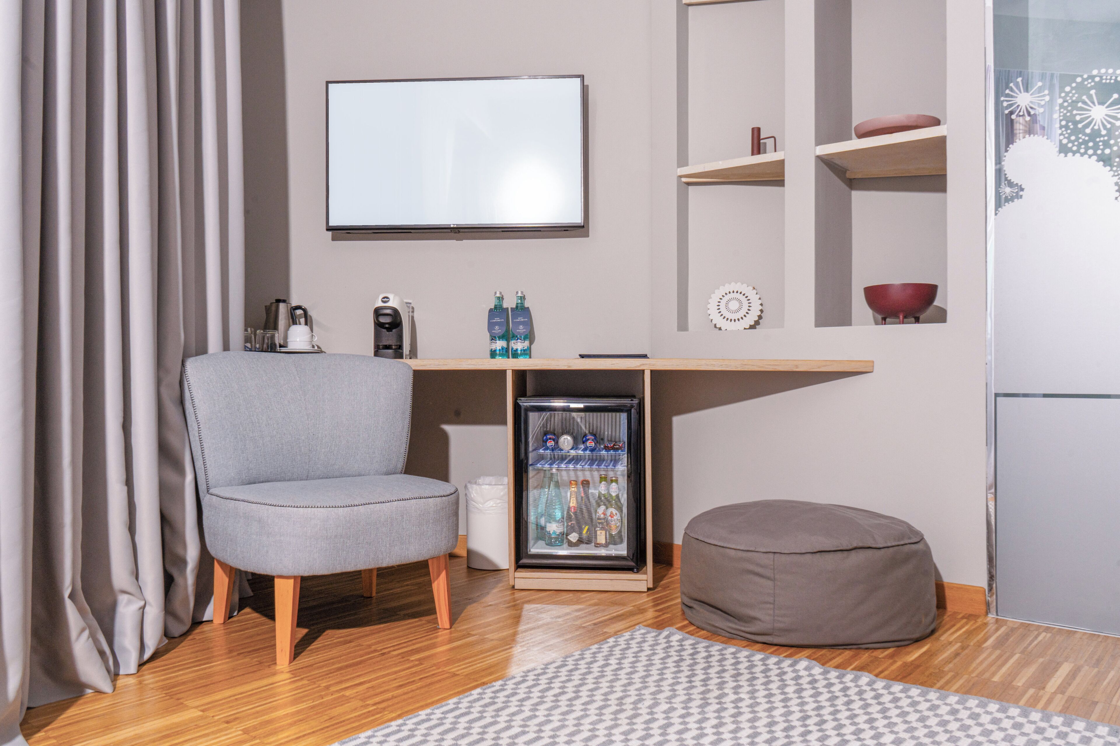 Een moderne kamer met een grijze fauteuil, een kleine koelkast, een houten bureau, een grijze pluchen zitbal en een kamerkleed met dessin.