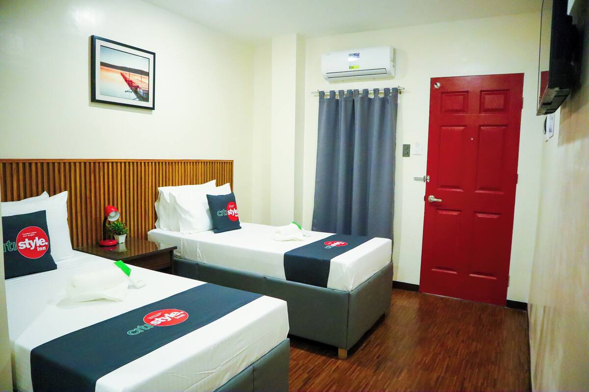Quarto de hotel com duas camas, uma porta vermelha, unidade de ar condicionado e um quadro na parede.