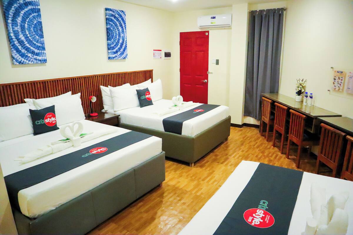 Quarto de hotel com duas camas, uma escrivaninha e uma cadeira. Há quadros nas paredes e uma porta vermelha.