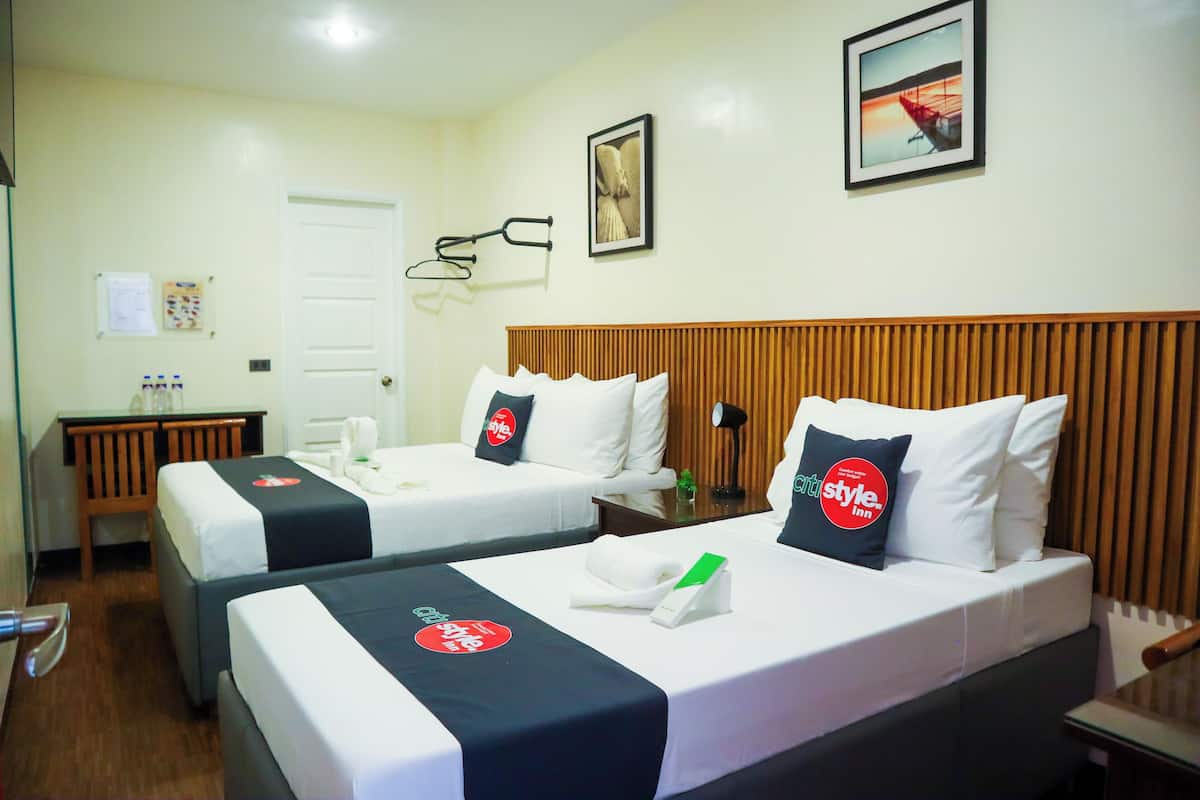 Quarto de hotel com duas camas, uma escrivaninha e uma cadeira. Há toalhas e uma placa verde nas camas.