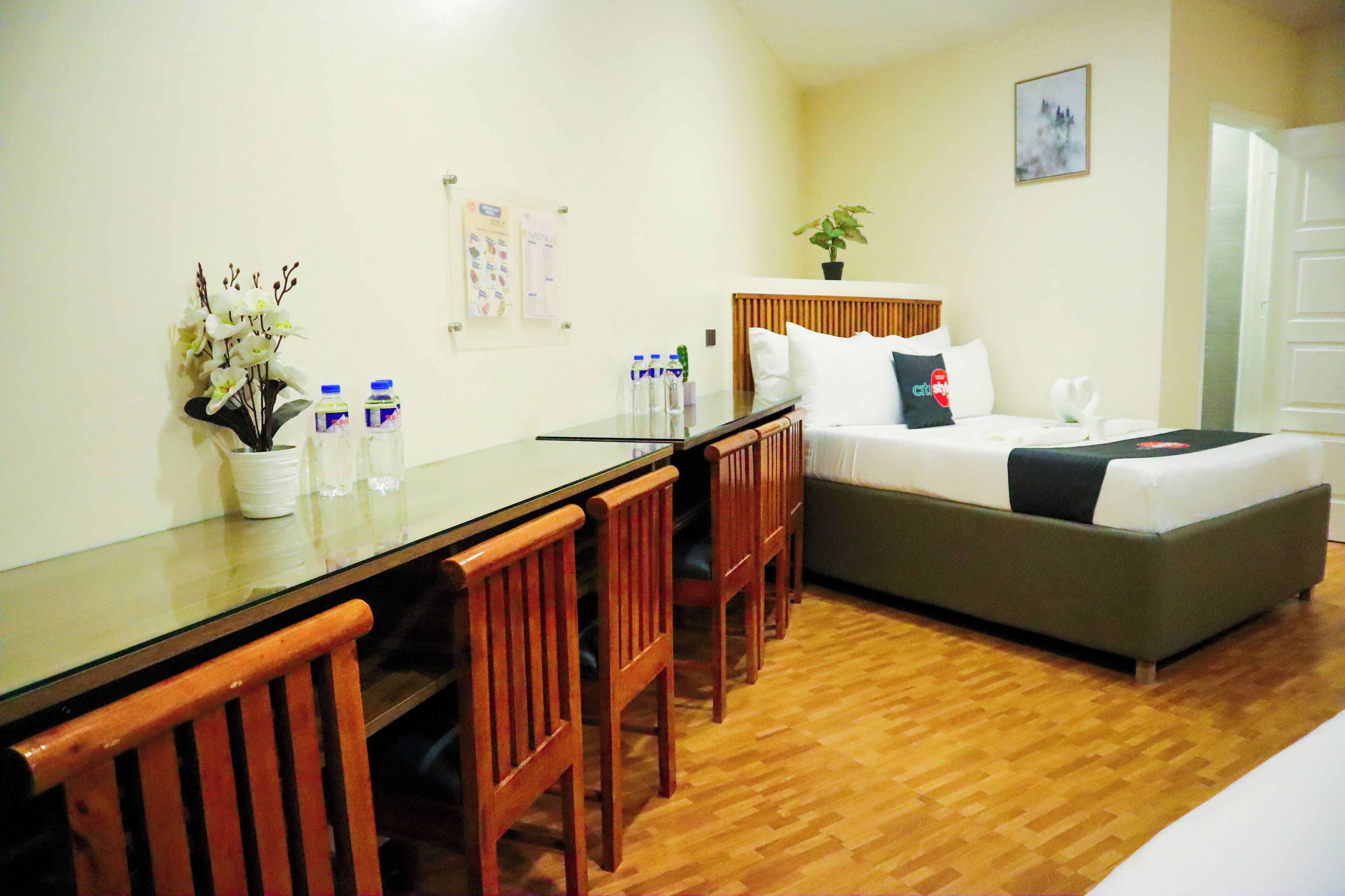 A hotel room with a wooden dining table, four wooden chairs, a bed with a green mattress, a wooden headboard, a potted plant, and a wall-mounted bulletin board.