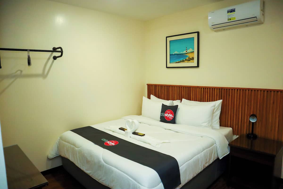 Quarto de hotel com uma cama bem arrumada, mesa de cabeceira com abajur, ar-condicionado fixado na parede e um quadro emoldurado acima da cama.