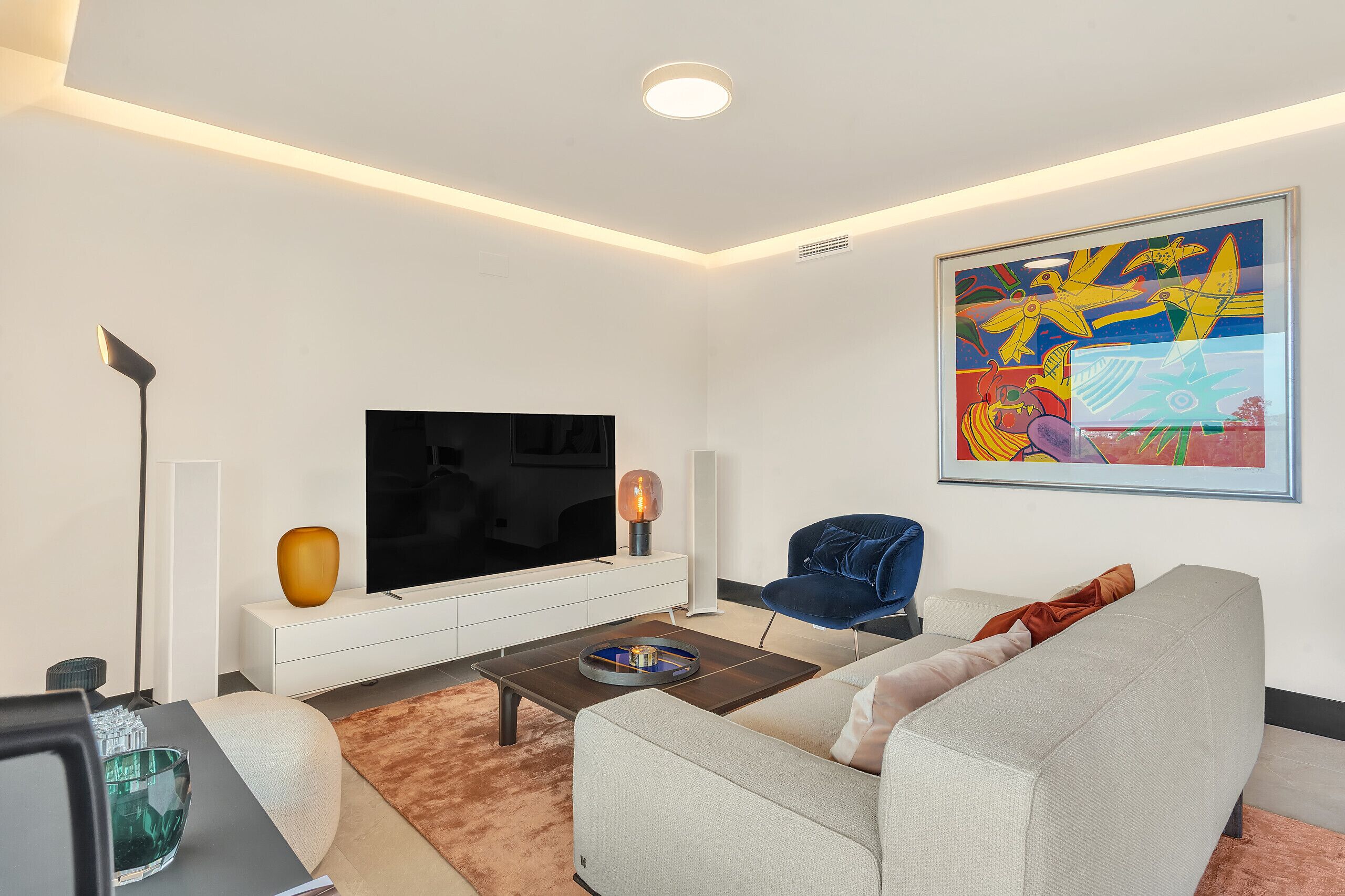 A modern living room with a flat-screen TV, a coffee table, a sofa, a blue chair, and a colorful abstract painting on the wall.