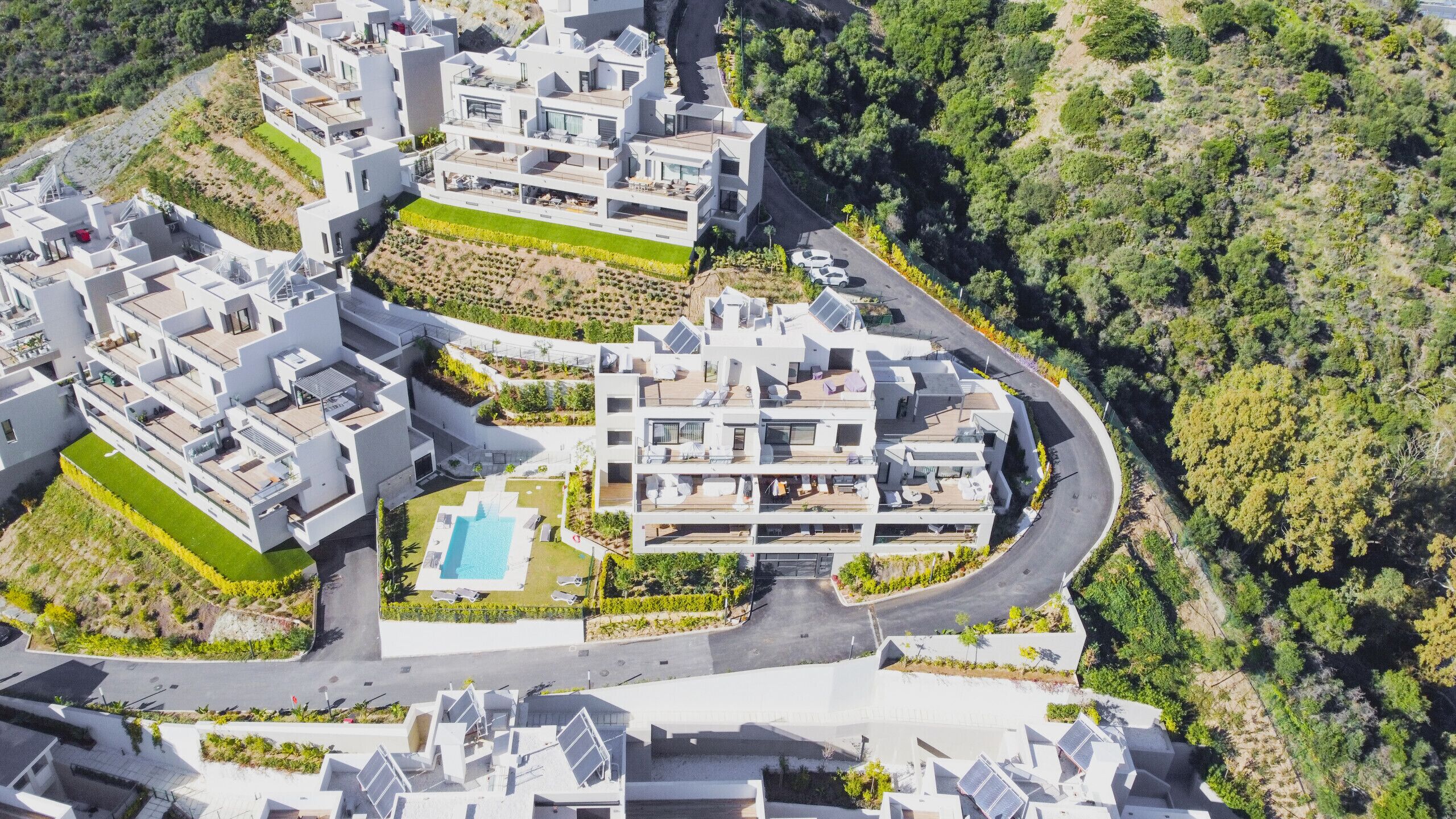 An aerial view of a modern residential complex with multiple buildings, a central swimming pool, and surrounding greenery.