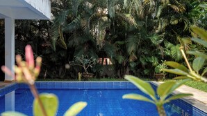 A swimming pool with a tiled edge, surrounded by lush greenery and a white building structure.