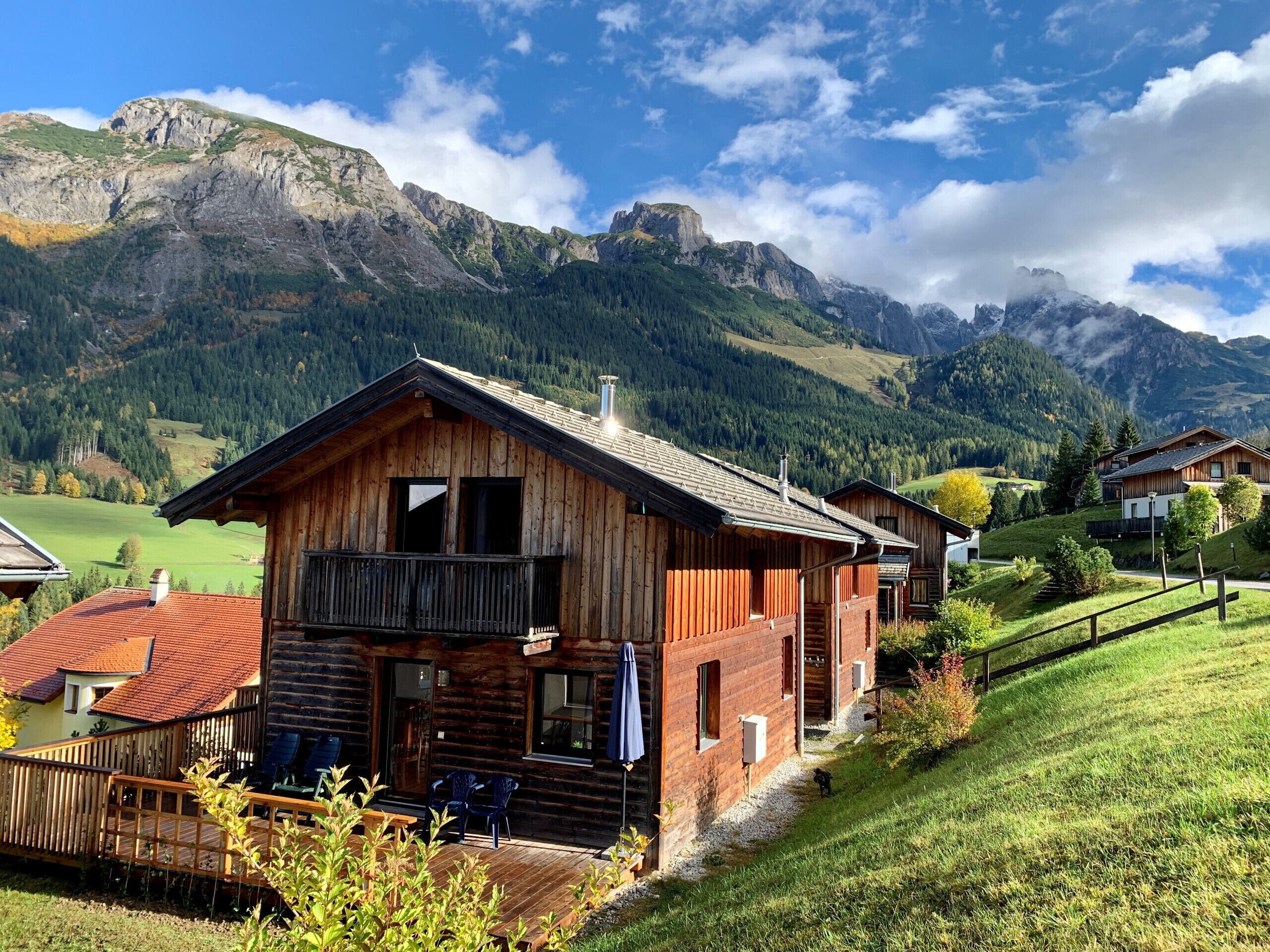 A wooden chalet with a balcony, surrounded by a lush green landscape and mountains in the background.