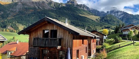 A wooden chalet with a balcony, surrounded by a lush green landscape and mountains in the background.