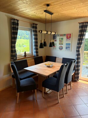 Uma sala de jantar com mesa de madeira, cadeiras pretas, luminária pendente e janela com cortinas.