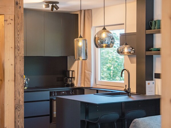A modern kitchen with a central island, pendant lights, and a view of greenery outside.