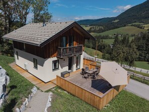 Un chalet moderne doté d’une terrasse en bois, de grandes fenêtres et d’un balcon offrant une vue imprenable sur une vallée pittoresque.