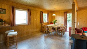 A wooden cabin interior with a dining table, chairs, a bench, a stove, and a window with curtains.