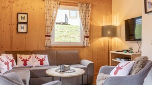 A cozy room with a sofa, armchair, and a small table. There is a window with curtains, a lamp, and a television on the wall. - Chalet # 92 with 3 BR for up to 6 people (Annaberg)