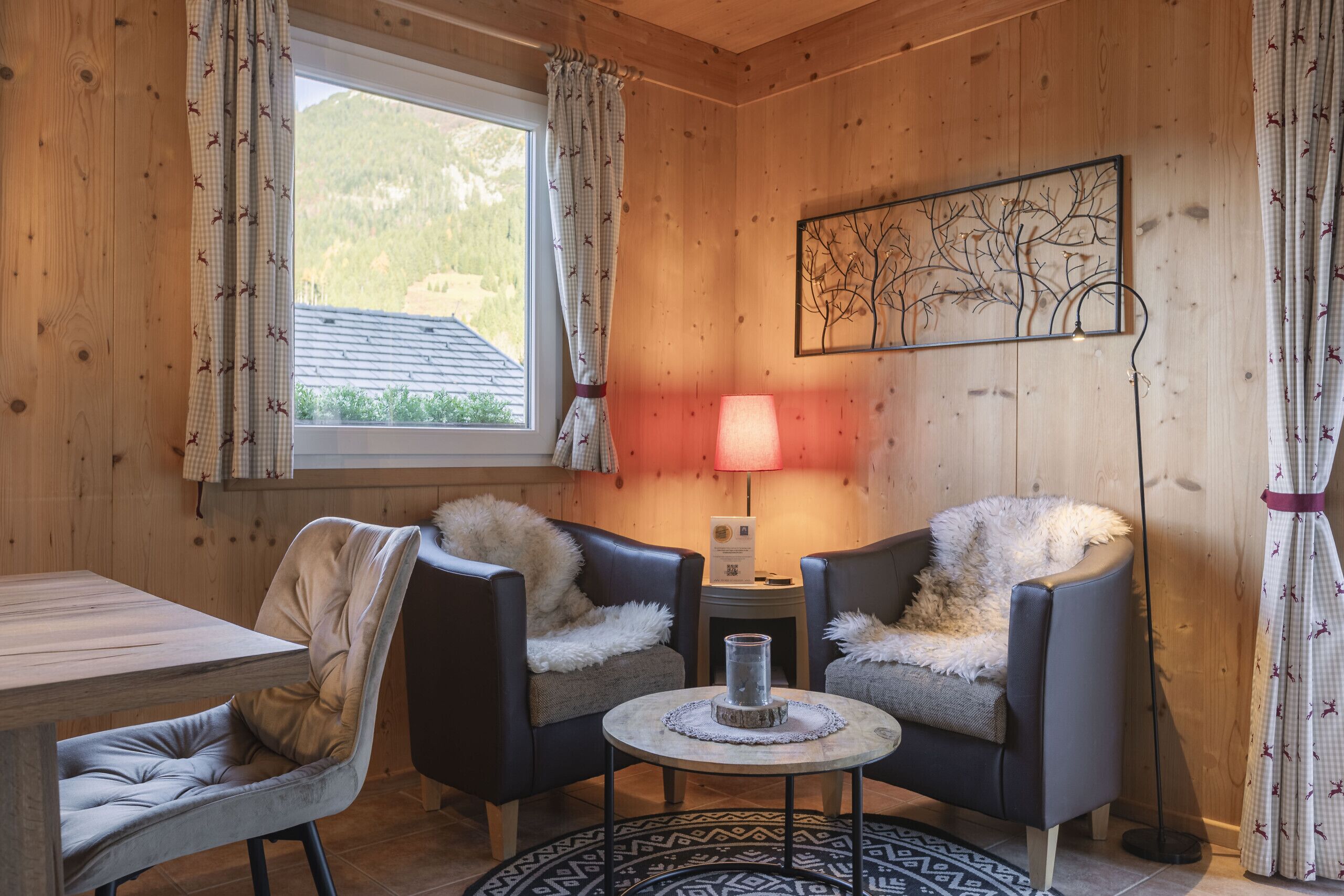 A cozy room with wooden walls, a dining table, two armchairs, a small side table with a lamp, and a framed artwork on the wall.