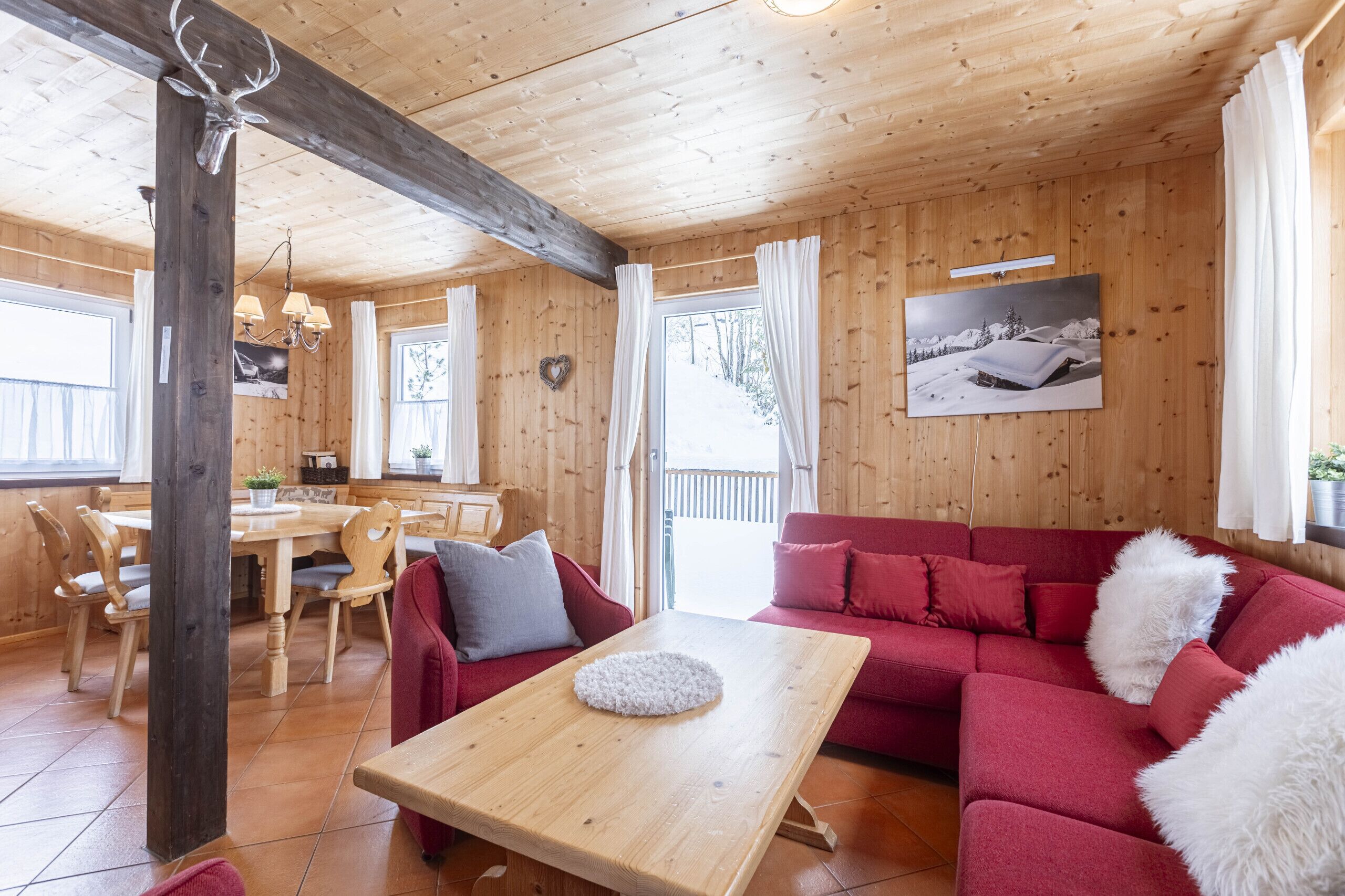 A cozy cabin interior with a wooden dining table, red sofa, and a large window with curtains.