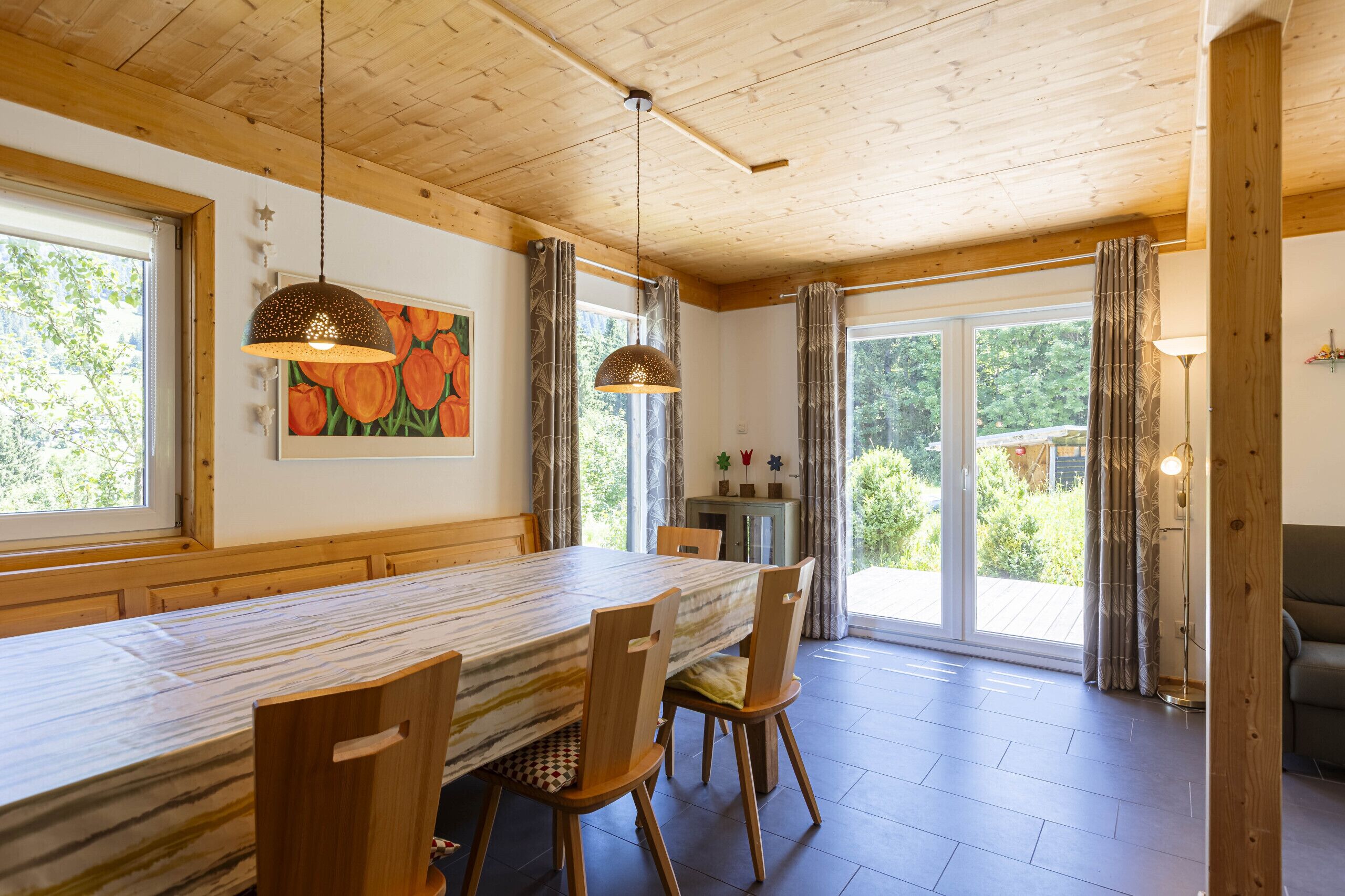 A dining area with a long table, wooden chairs, pendant lights, and a view through glass doors to the outside.