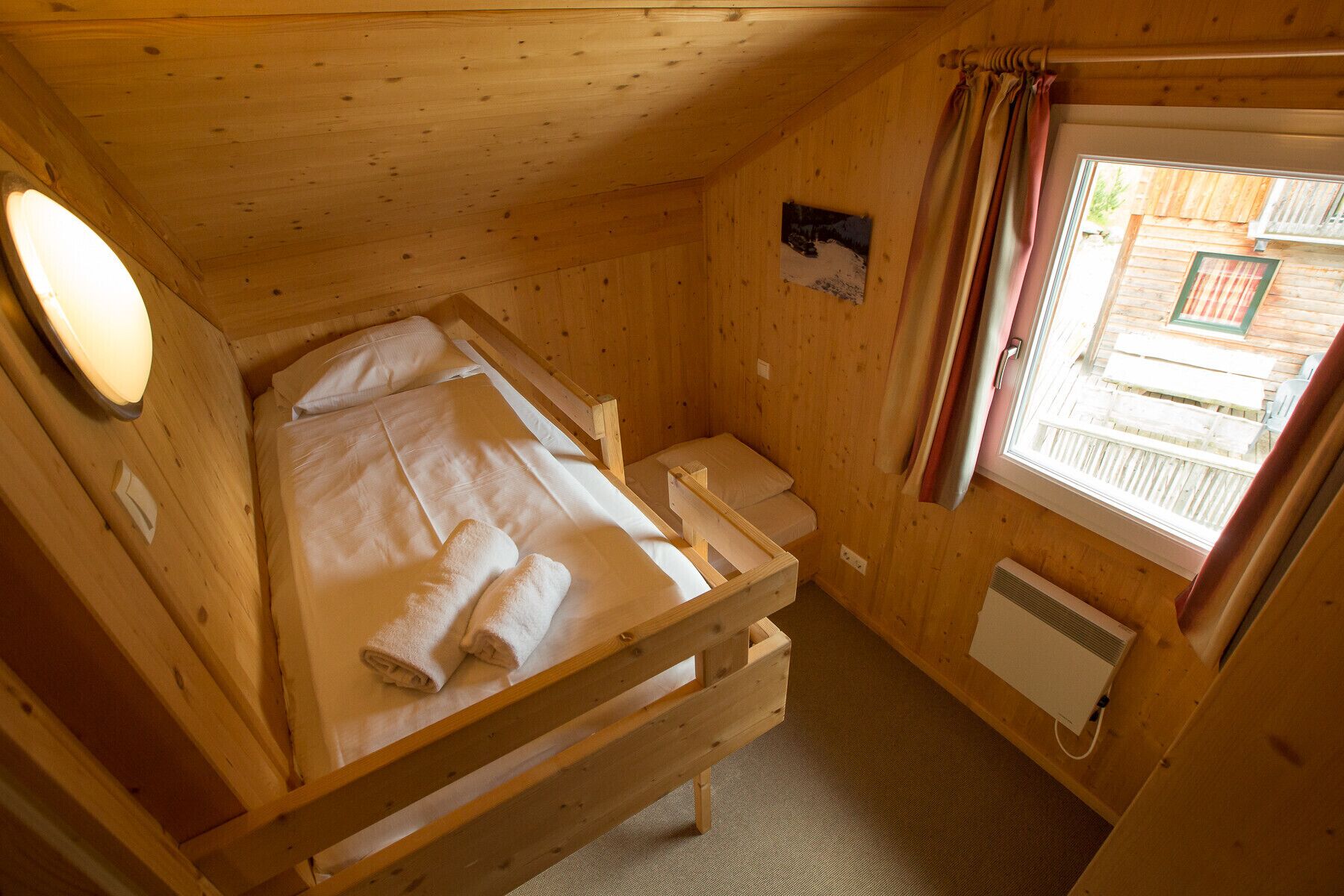 A small wooden cabin room with a bunk bed, a single bed, a window with curtains, a heater, and a wall-mounted light.