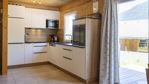 Una cocina moderna con armarios blancos, un microondas de acero inoxidable y un refrigerador grande. Se ve una ventana con vistas a una terraza de madera y montañas.