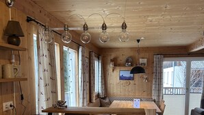 L’intérieur d’un chalet en bois accueillant présente un espace cuisine avec une table en bois, des chaises et un plan de travail noir. On y trouve plusieurs fenêtres avec des rideaux, un plafond orné de luminaires suspendus et une vue sur l’extérieur à travers la porte vitrée.