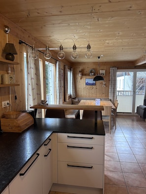 L’intérieur d’un chalet en bois accueillant présente un espace cuisine avec une table en bois, des chaises et un plan de travail noir. On y trouve plusieurs fenêtres avec des rideaux, un plafond orné de luminaires suspendus et une vue sur l’extérieur à travers la porte vitrée.
